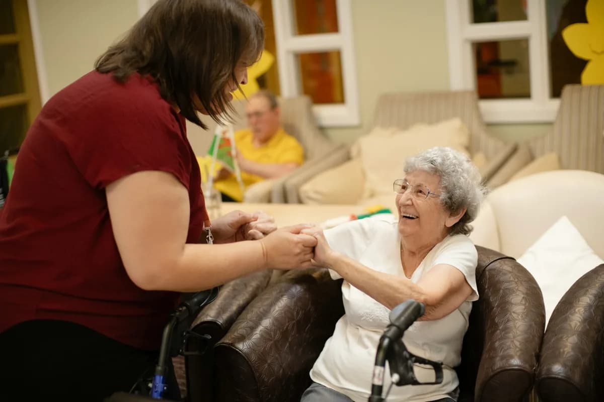 Soignante tenant les mains d’une résidente souriante dans le salon d’une maison de retraite